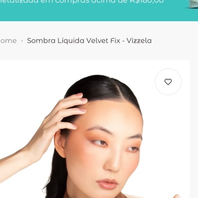 Modelo de cabelo preto usa blusa marrom, toca o rosto e segura embalagem de maquiagem diante de fundo branco