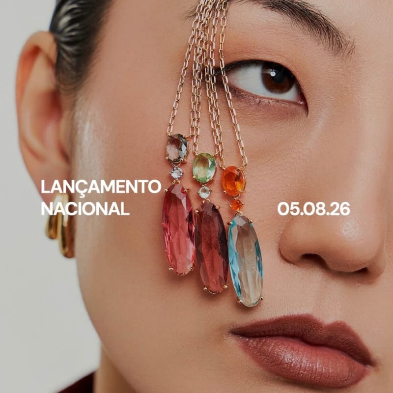 Close de modelo com cabelo preso, correntes douradas e pingentes coloridos sobre o rosto, em fundo neutro
