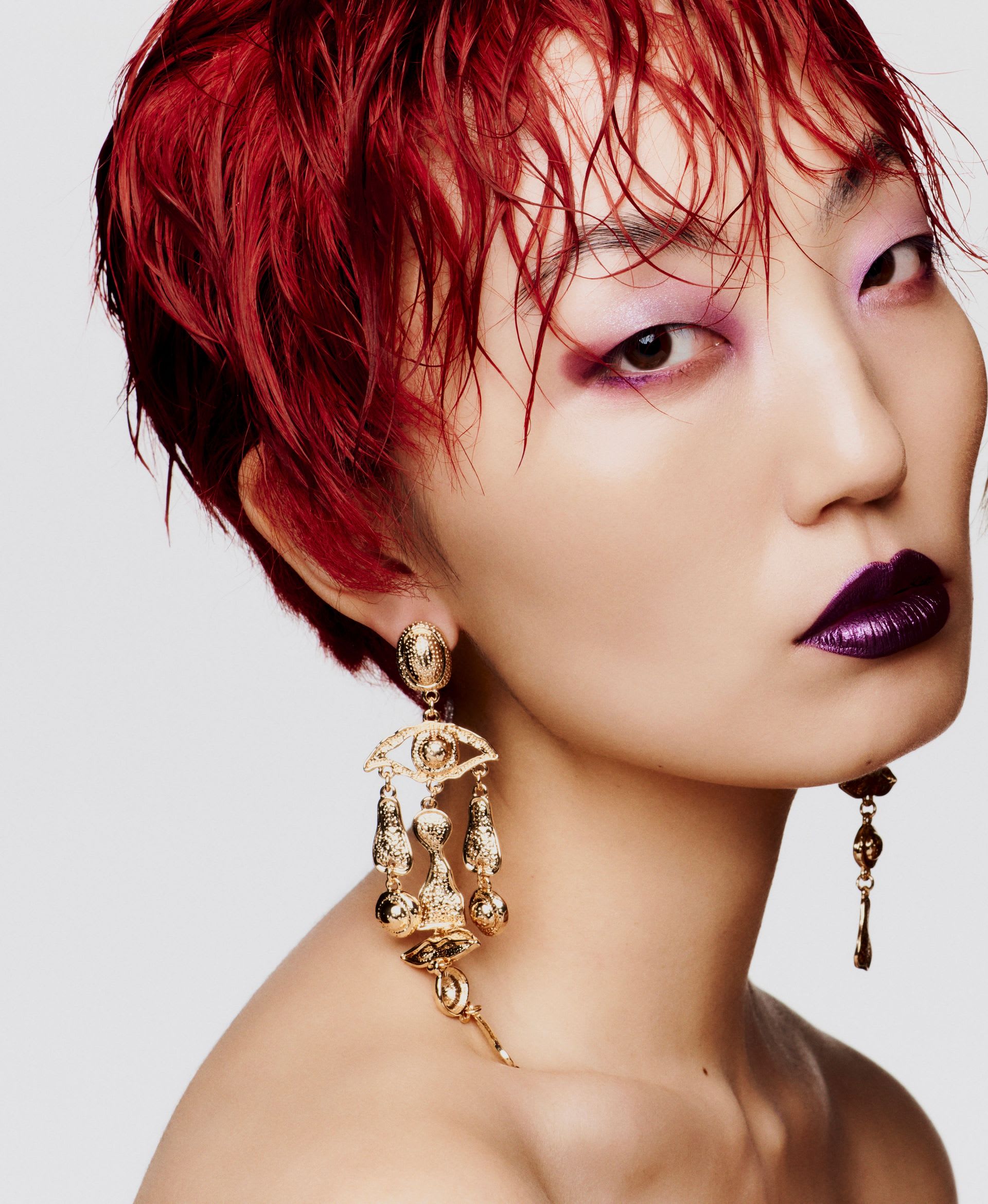 Modelo com cabelo curto vermelho molhado, batom roxo e brincos dourados, em close e de perfil contra fundo claro