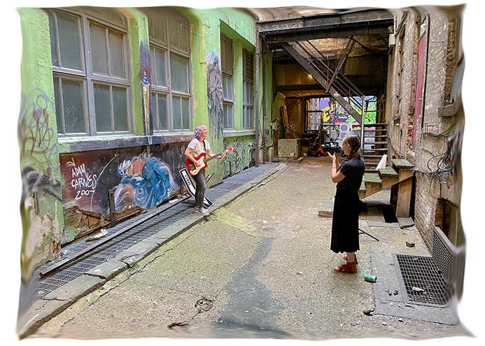 color photo in a graffiti'd alley with Elyza playing guitar and singing while being filmed