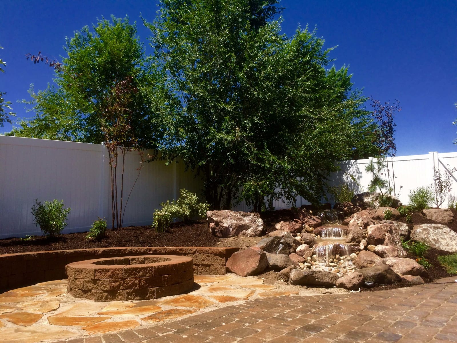 Stone patio with a circular fire pit, boulder waterfall, and established trees