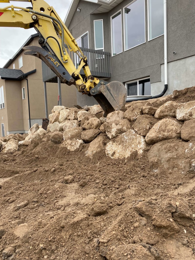 Excavator placing large boulders for a retaining wall beside a house
