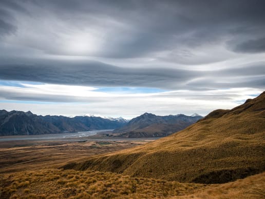 New Zealand Alpine Hikes, Trekking & Snowshoeing - New Zealand Alpine Treks & Hikes - Tekapo High Country Hike - Tekapo Trek - views of the Southern Alps (photo: K. Bowie) - A vast, rugged landscape with towering mountains in the background, a dry, grassy foreground, and a dramatic, cloudy sky overhead. 4