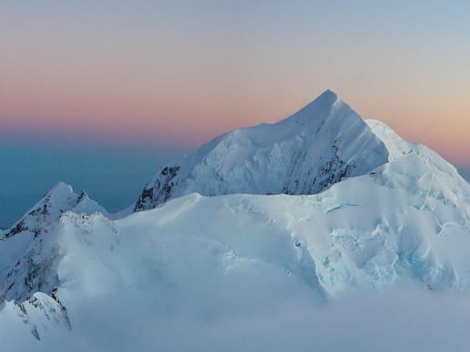 New Zealand Alpine Climbing, Mountaineering Instruction & Guided Ascents - Guided Alpine Ascents in New Zealand - Mount Tasman - Mount Tasman - sunrise - A majestic, snow-capped mountain peak rises above a sea of clouds, bathed in the warm glow of a sunset sky. 1