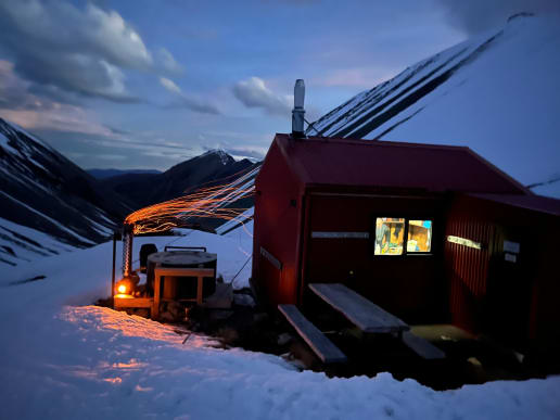 New Zealand Backcountry Skiing & Split-boarding - The Best Freeride, Ski Touring & Split-Boarding Adventures in New Zealand - Cass Valley Ski & Split-board Tours - O'Leary's Hut and hot tub at the head of Waterfall Stream, Cass Valley Ski Touring - A red mountain hut with a lit window sits in the snowy foreground, while a wood fire with glowing sparks and a smoky chimney heats a hot tub to its left, all set against a twilight sky and snow-covered mountains in the background. 3