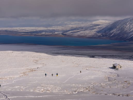 New Zealand Alpine Hikes, Trekking & Snowshoeing - New Zealand Snowshoe Hikes & Winter Treks - Lake Tekapo Snowshoeing - Snowshoers returning to Rex Simpson Hut above Lake Tekapo, Two Thumb Range, New Zealand - A snowy, mountainous landscape with a vibrant blue lake in the background, and several people walking across the snow-covered foreground. 4