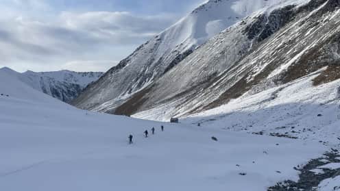 Cass Valley Falcon's Nest Hut | Ski touring and Split-boarding adventures in the Gamack Range - A snowy, mountainous landscape with a group of people trekking across the frozen terrain in the foreground, surrounded by towering peaks and a cloudy sky in the background. 1