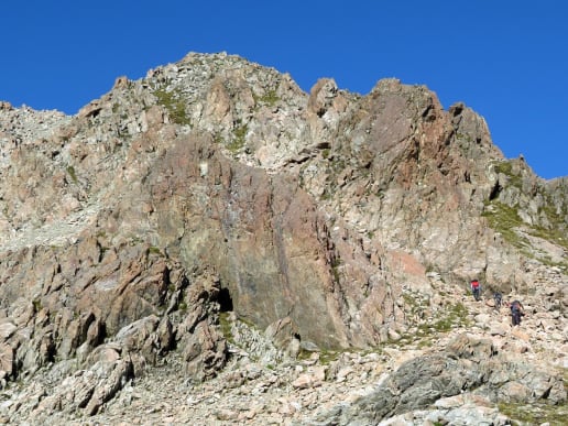 About - Preparing for your Mountain Adventure - Routes to and from Caroline Hut and Ball Ridge - Hikers climbing up untracked rocky alpine terrain above Caroline Hut on the way to Ball Ridge - Rugged, rocky mountain peaks rise against a clear blue sky, with a steep, craggy foreground leading up to the towering formations. 4