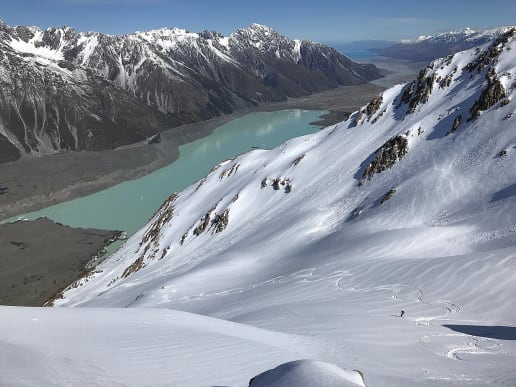 New Zealand Backcountry Skiing & Split-boarding - New Zealand's Best Glacier Touring & Ski Mountaineering - Ski Mount Cook Range - Skiing down from Fergun's Knob near Caroline Hut, in front of the Tasman Glacier Lake, Aoraki Mount Cook National Park - A breathtaking snowy landscape with towering mountains, a turquoise glacial lake, and a lone skier navigating the powdery slopes in the foreground. 1