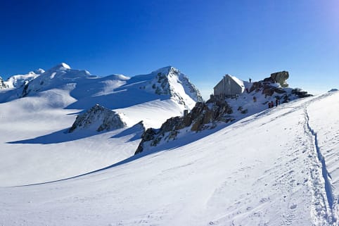 Venue: Kelman Hut (winter) - A snow-covered mountain landscape with towering peaks and a winding trail leading through the snowy terrain under a clear blue sky.