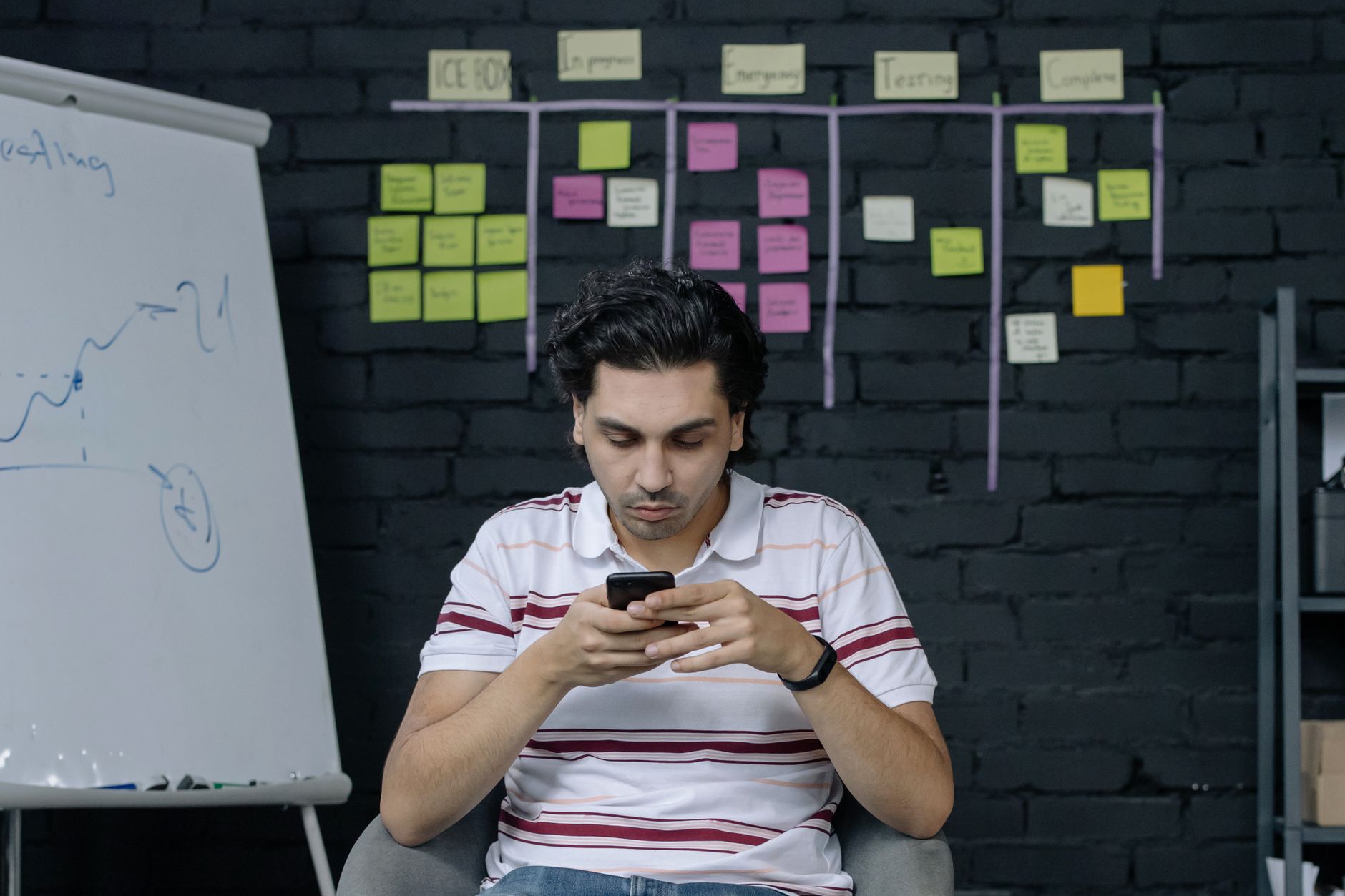 Man in office using smartphone against a black brick wall with sticky notes and whiteboard.