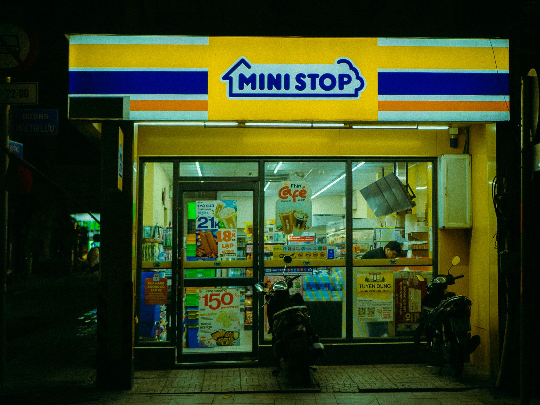 A brightly lit Mini Stop convenience store at night with motorbikes parked in front.