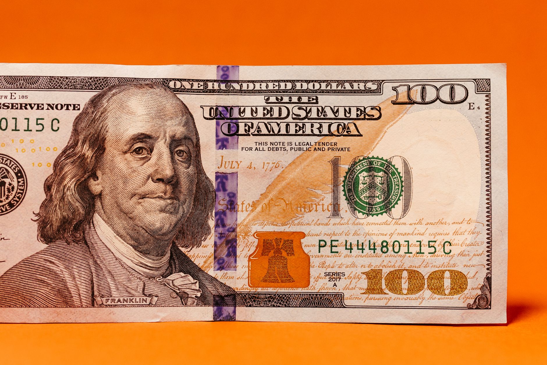 Detailed image of a US hundred dollar bill against a vibrant orange background.