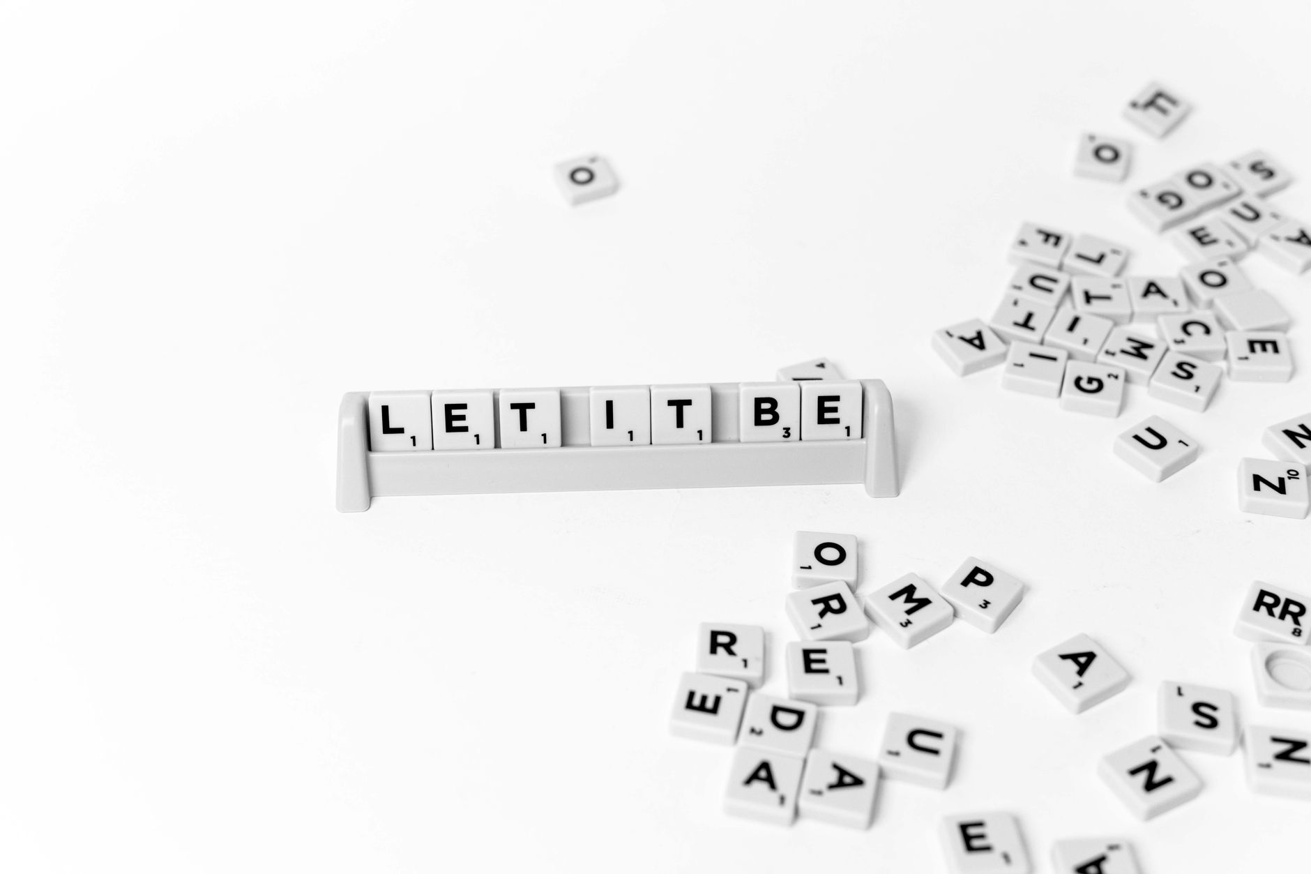 Flat lay of Scrabble tiles spelling 'Let It Be' with scattered letters on a white background.