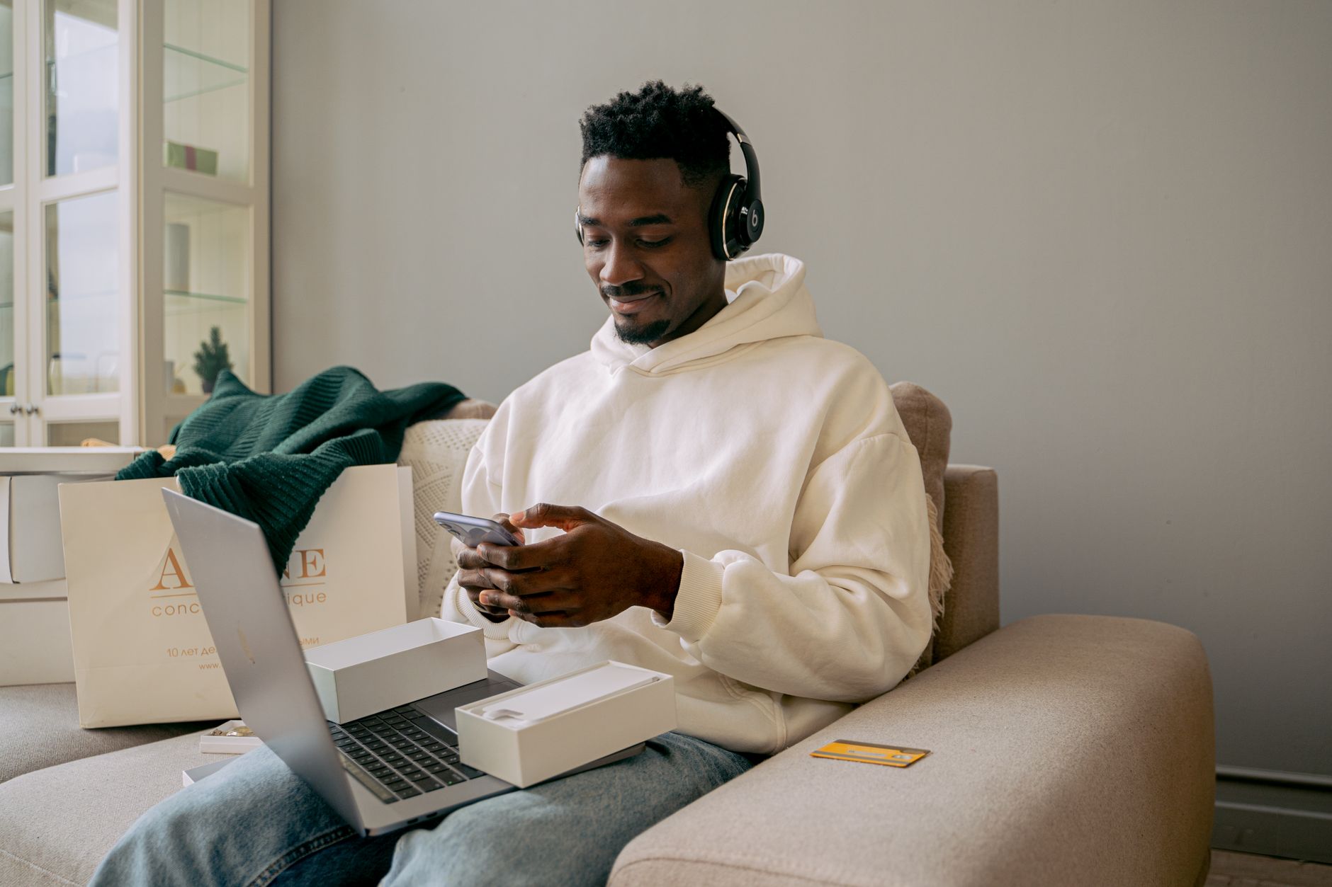 Man in a hoodie using a new smartphone and laptop on a sofa, indoors.