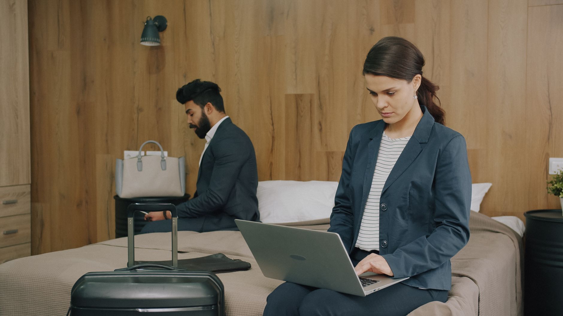 A man and woman in business attire working on laptops in a modern hotel room.