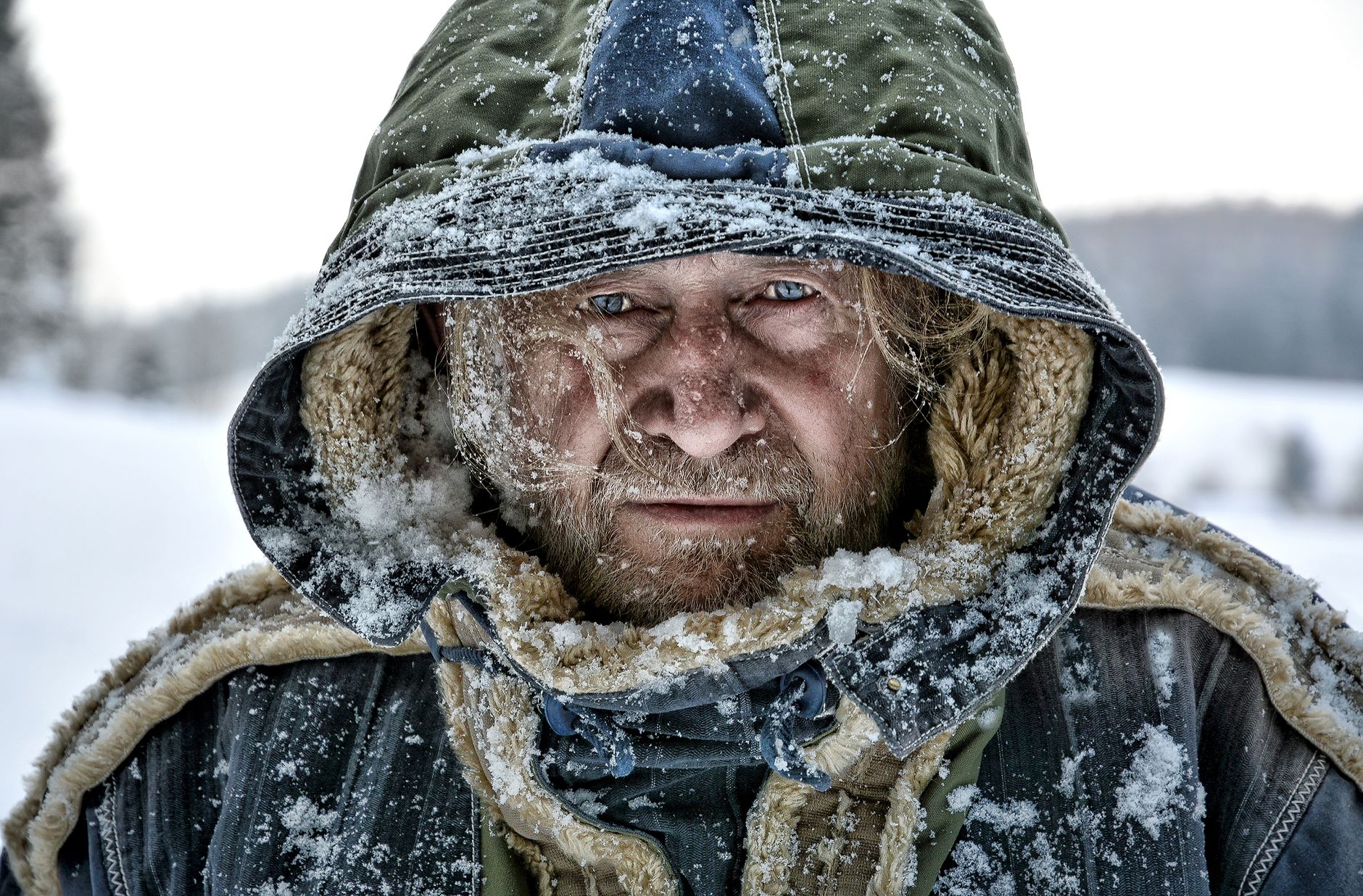 Close-up portrait of a mature man in a hooded jacket covered in snow, showcasing the harsh winter weather.