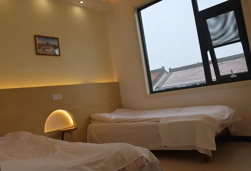 A clean, well-lit dormitory room in Wenxian County, Henan Province, featuring two beds, a bedside lamp, and a window overlooking traditional rooftops.