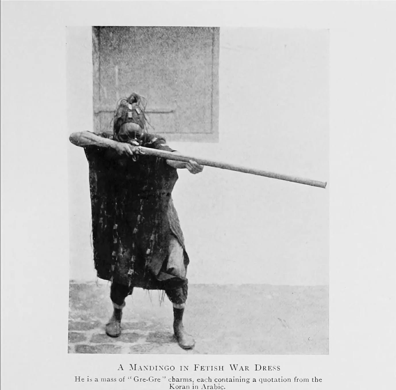 A Mandingo man in war dress covered in gris-gris, each holding a verse of the Koran, 1910