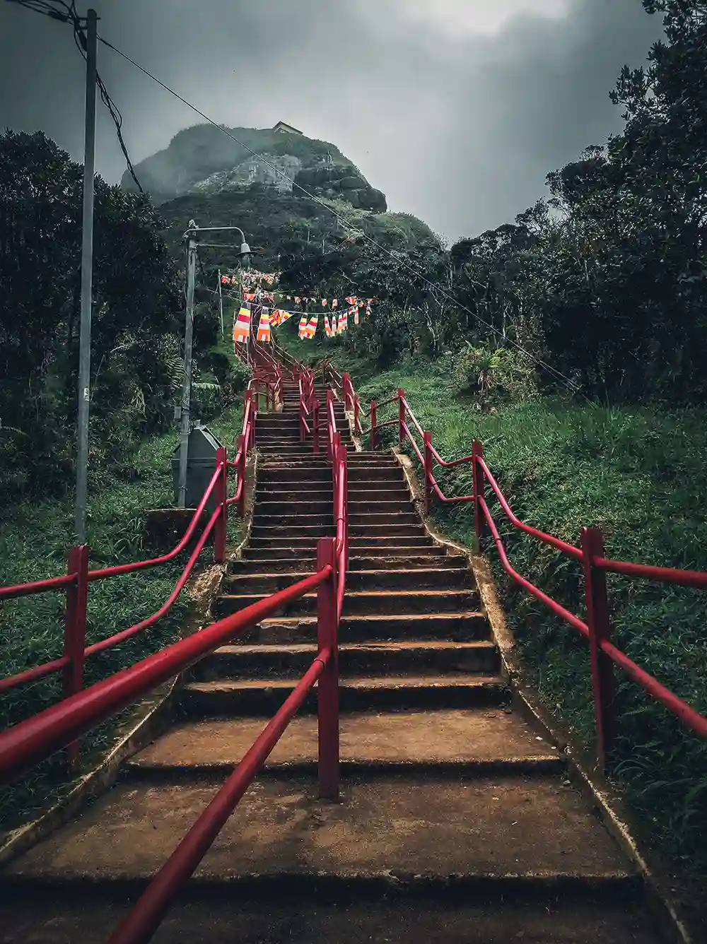 A low-angle view of the steep stone steps on the Adam's Peak trail, lined with red railings and decorated with Buddhist prayer flags.