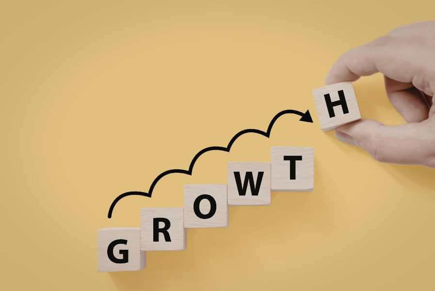 A hand places a wooden block with the letter "H" at the end of a row spelling "GROWTH" on a yellow background.