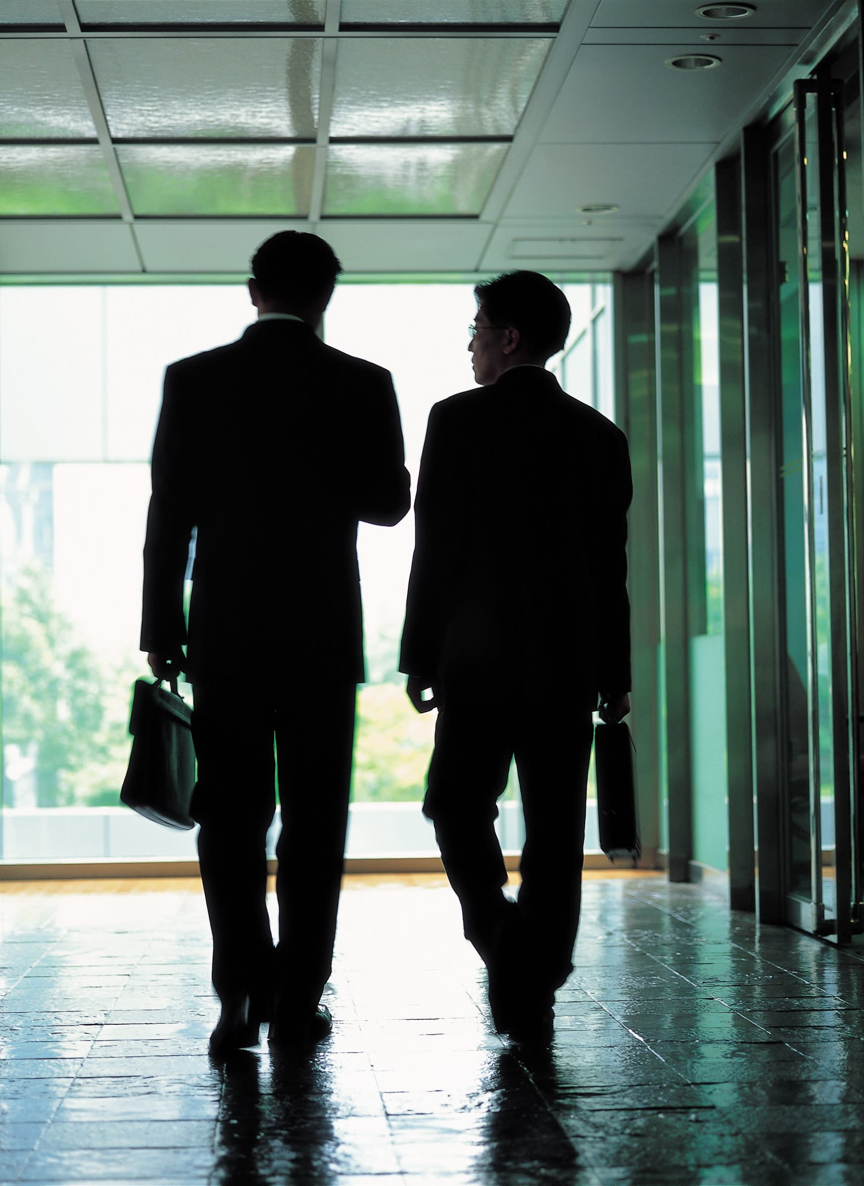 2 men holding briefcases and walking down a hallway talking with one another