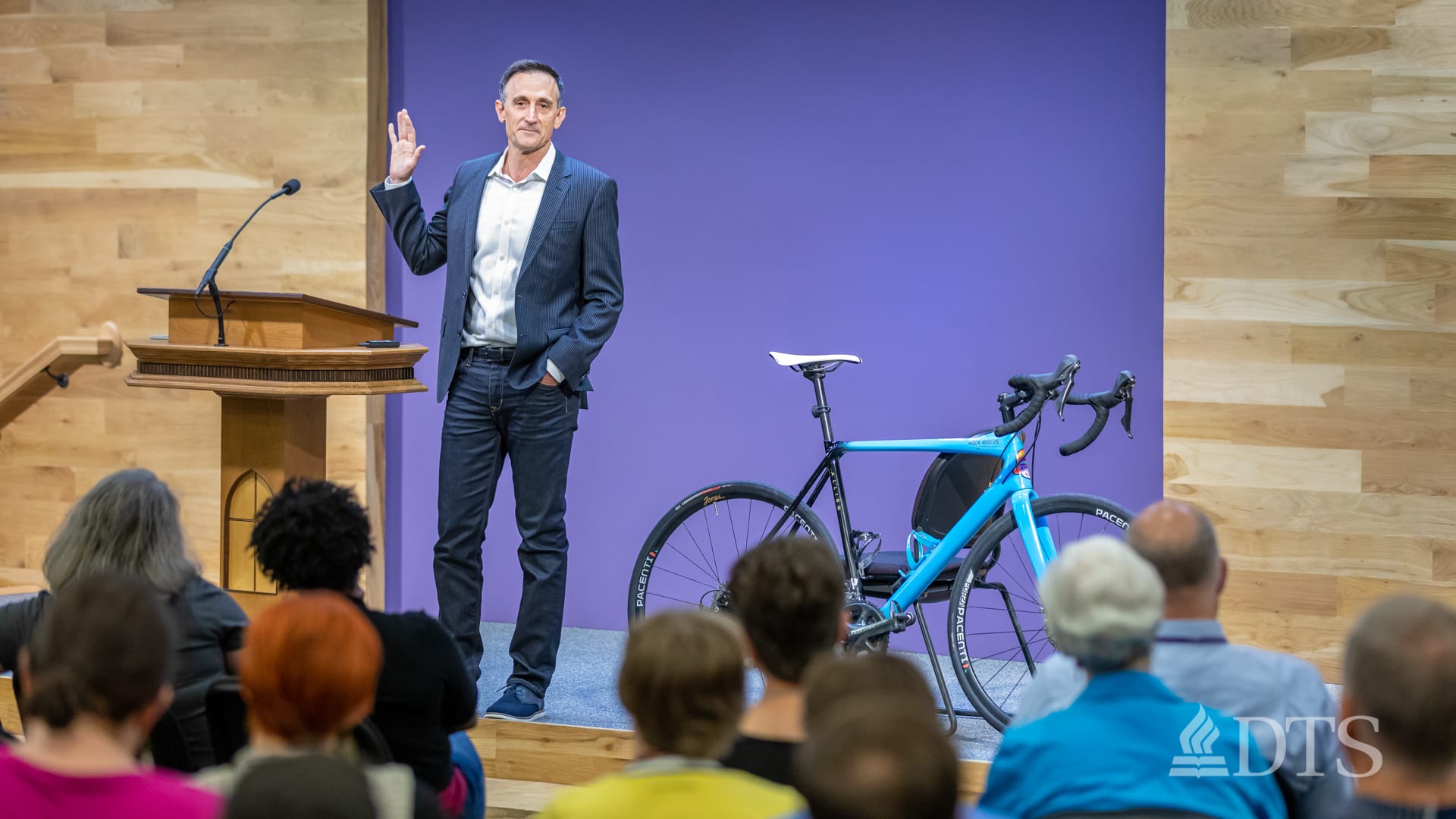 man standing next to podium with bicycle on stage speaking at DTS Chapel