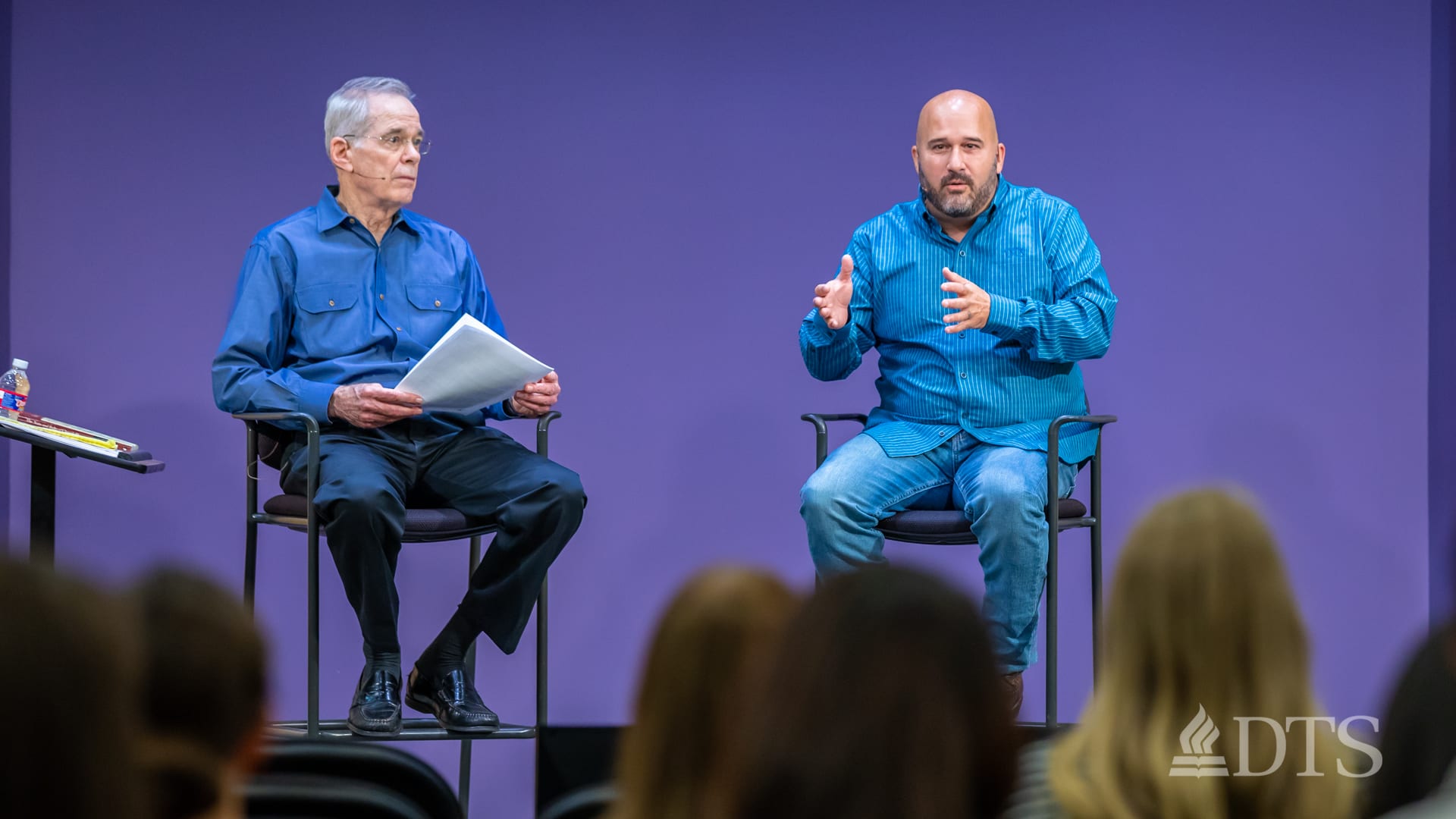 two men seated on stools on stage while speaking at DTS Chapel
