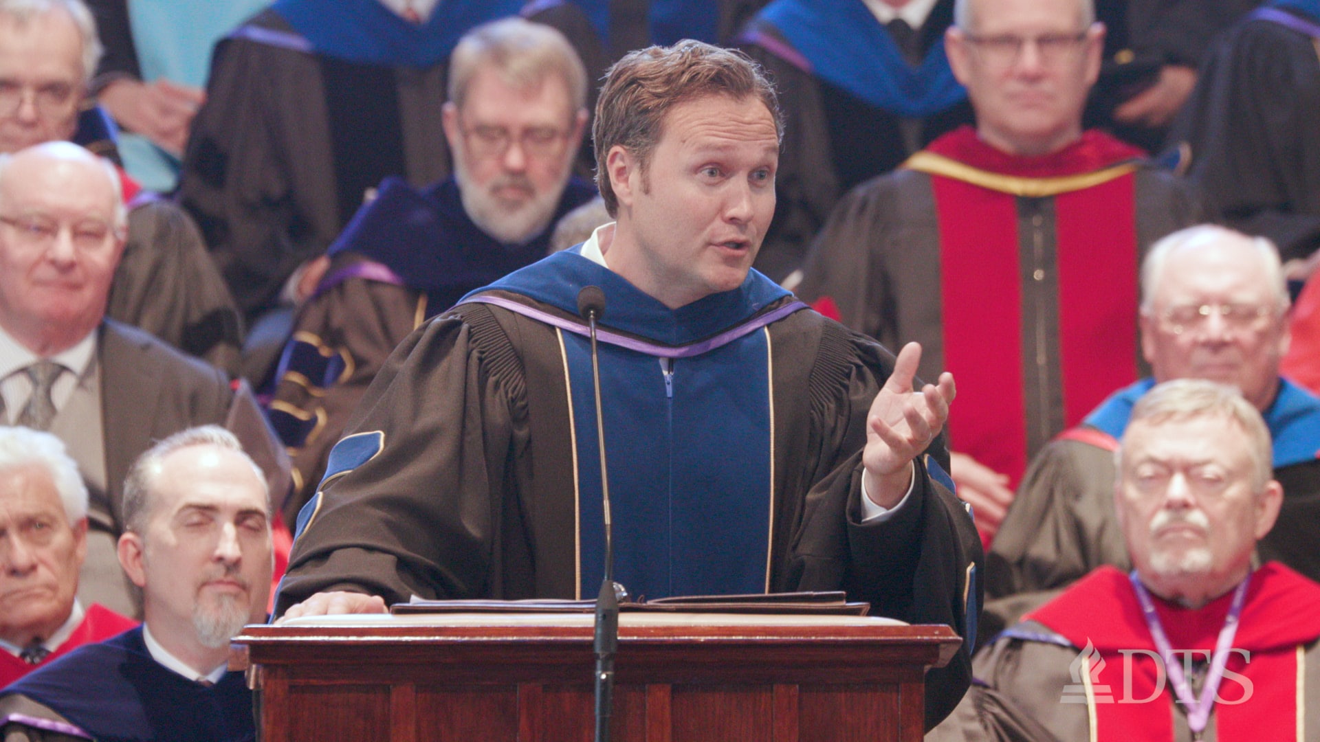 man standing at podium in regalia leading prayer at DTS Commencement Chapel