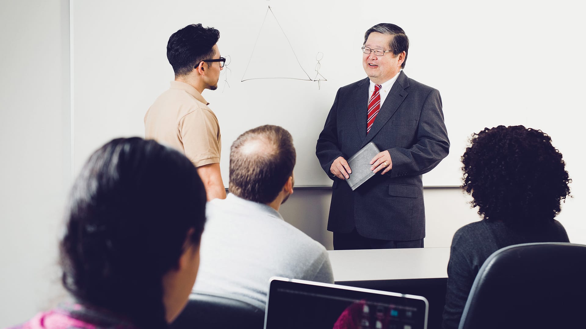 2 men standing together talking in front of a classroom full of students
