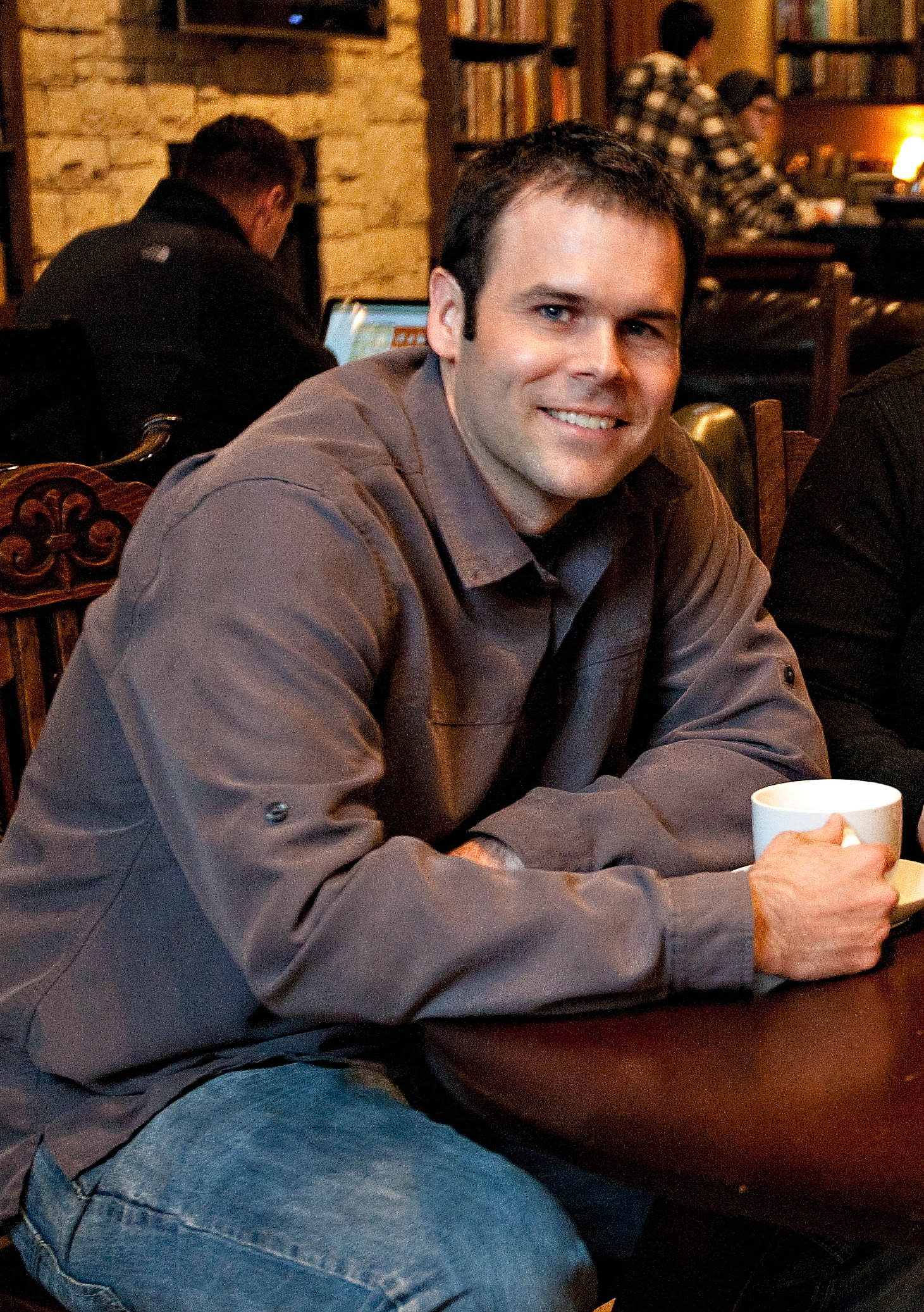 Man seated at a table holding a coffee cup and smiling while looking at the camera