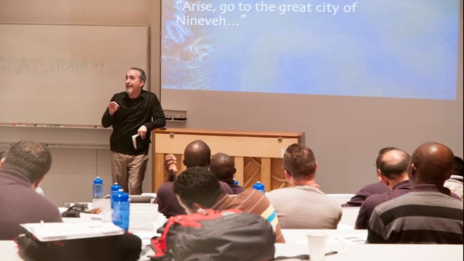 Dr. Mark Yarbrough leaning against piano teaching in a DTS classroom full of students