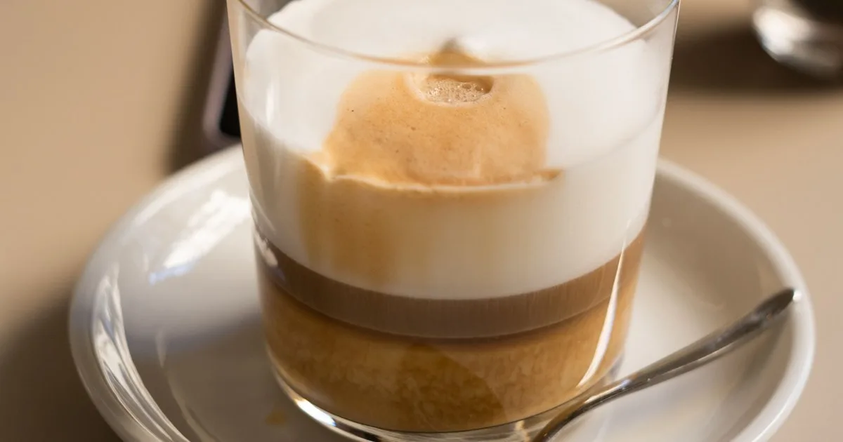 A latte macchiato in a glass tumbler showing three distinct layers: white foam on top, a band of espresso through the middle, and steamed milk at the bottom
