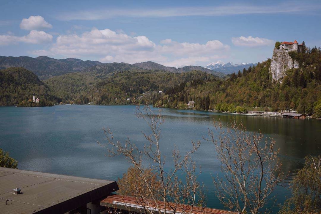 A scenic view of Lake Bled with its iconic island church and castle on a hill, surrounded by lush green forests and distant mountains under a partly cloudy sky.