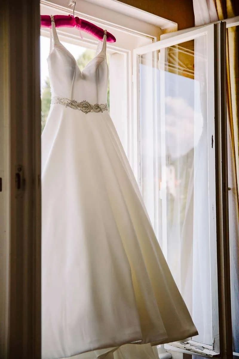A white wedding dress hanging on a pink hanger by an open window, allowing natural light to illuminate its elegant design.