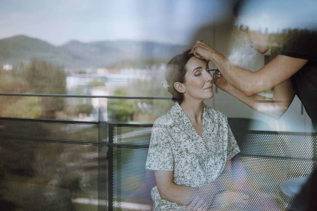 A bride sitting calmly as a makeup artist applies eyeshadow, with a scenic view outside the window.
