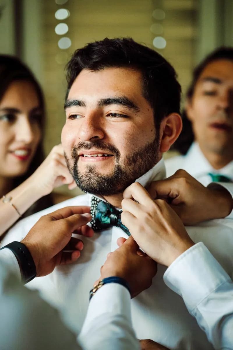 A groom smiling as his friends help adjust his bowtie before the wedding ceremony.
