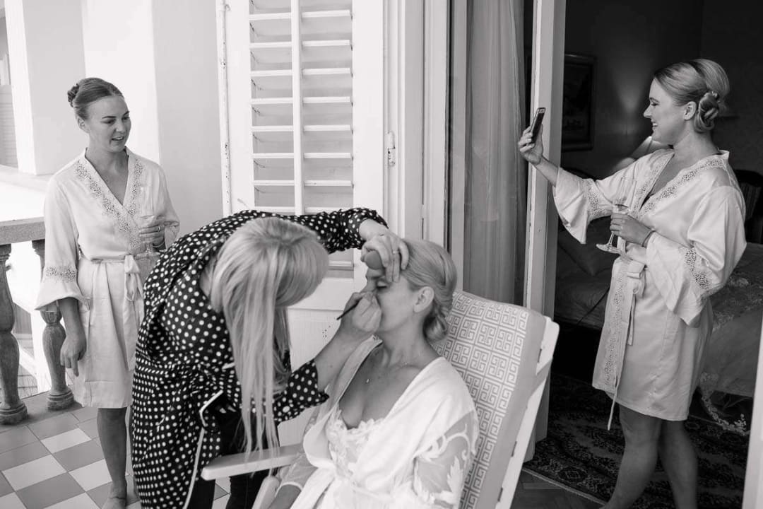 A bride having her makeup applied by an artist while two bridesmaids, wearing robes, look on.