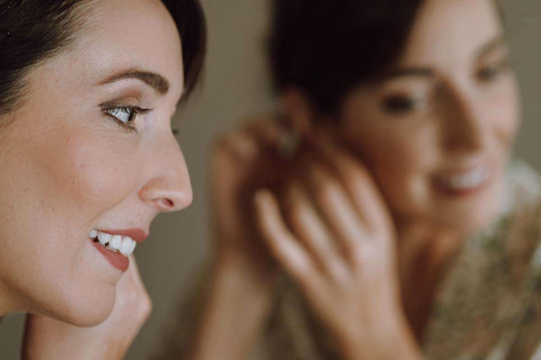A bride smiles as she puts on earrings, her reflection visible in a mirror.