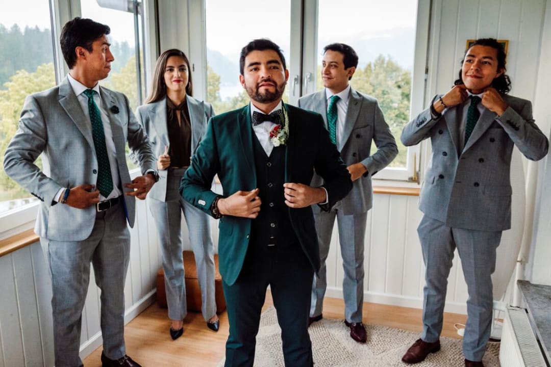 A groom in a dark suit stands confidently with his groomsmen, who are adjusting their suits, inside a bright room with a view of trees outside the window.