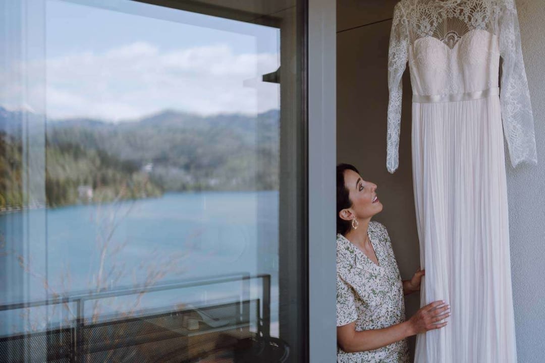 A bride admiring her lace wedding dress by a window with a view of Lake Bled in the background.