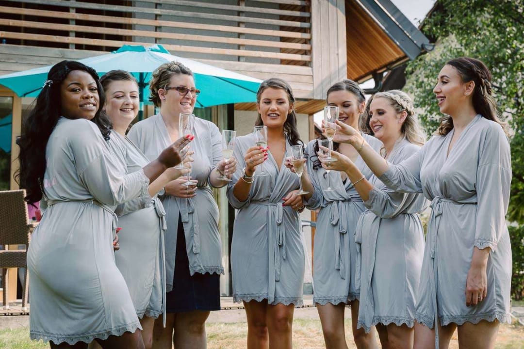 A group of bridesmaids in matching robes raising glasses in a toast outside a building, enjoying a wedding celebration.