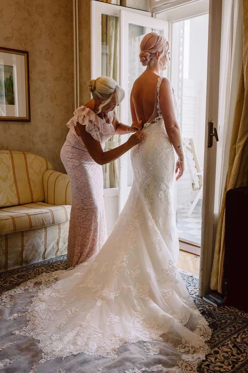 A bride standing in a room while an older woman helps adjust her lace wedding gown.