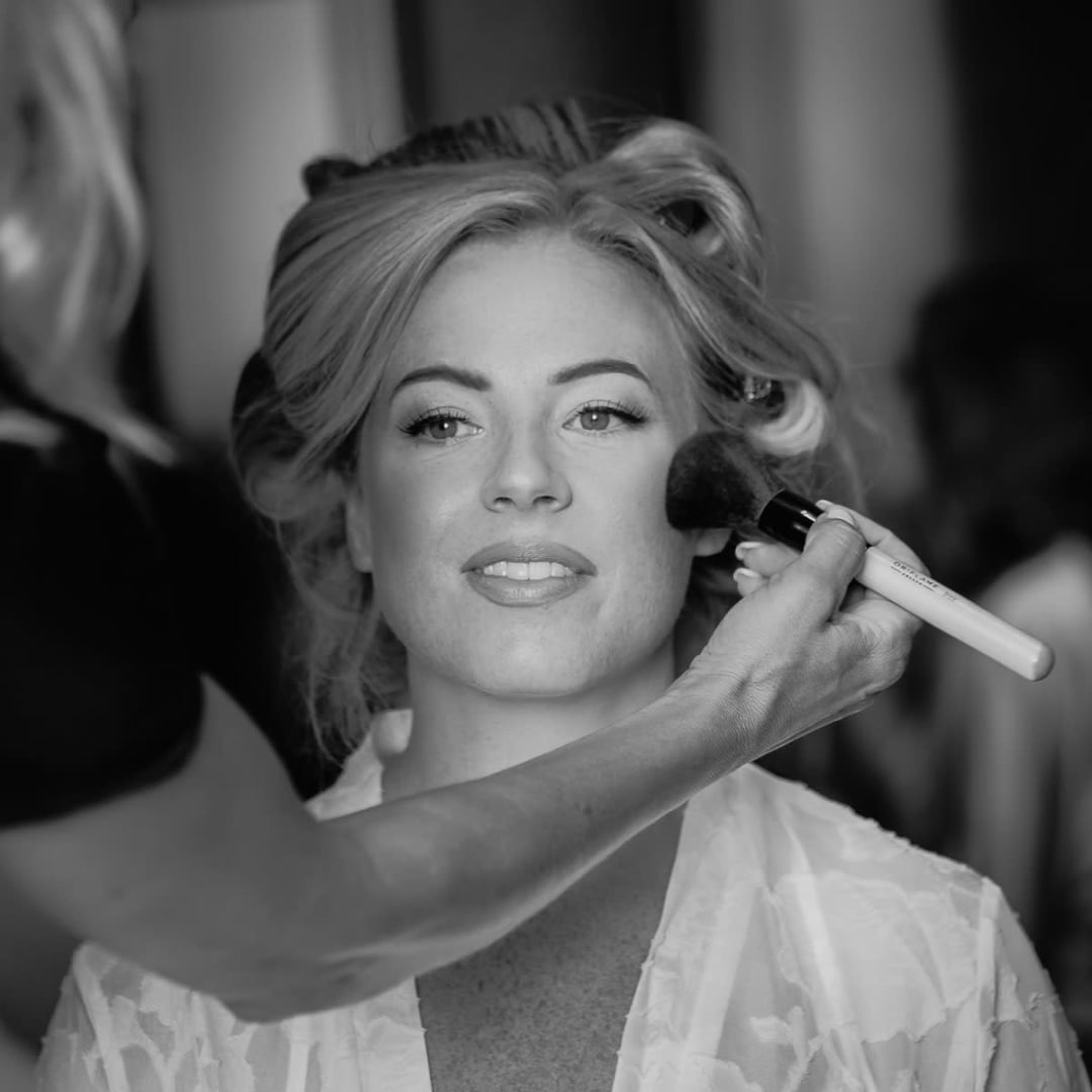 A bride getting her makeup done, smiling softly as a makeup artist applies blush to her cheek.