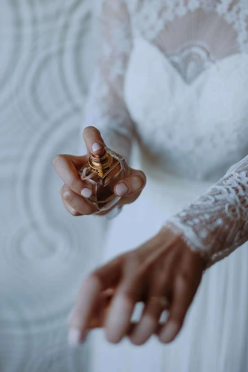 A close-up of a bride in a lace wedding dress applying perfume to her wrist.
