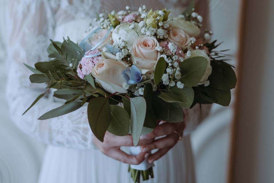 A bride in a lace dress holding a bouquet of roses and greenery in her hands.