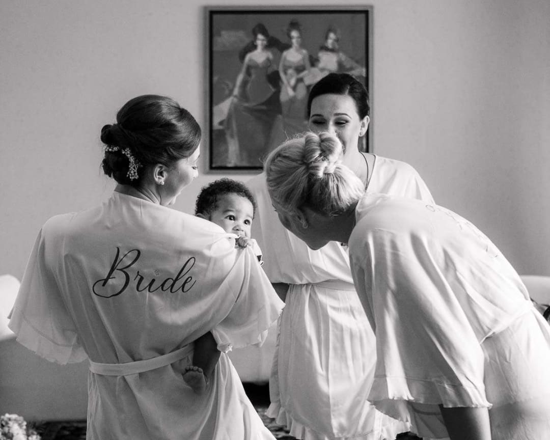 A bride and her friends in matching robes share a joyful moment with a baby before the wedding ceremony.
