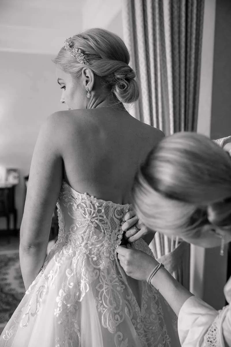 A bride has her intricate lace wedding dress fastened by a bridesmaid in a softly lit room.