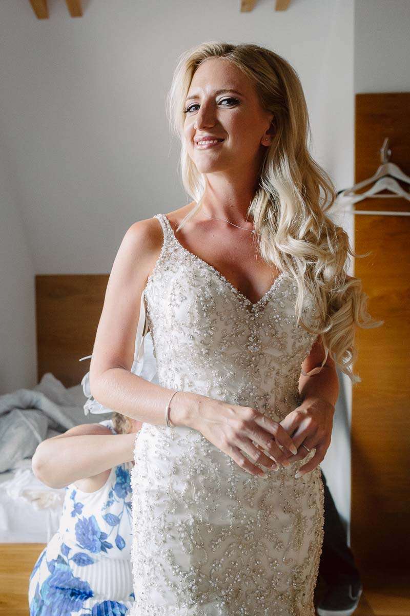 A bride smiling while getting ready, wearing a detailed white wedding dress as someone assists her.