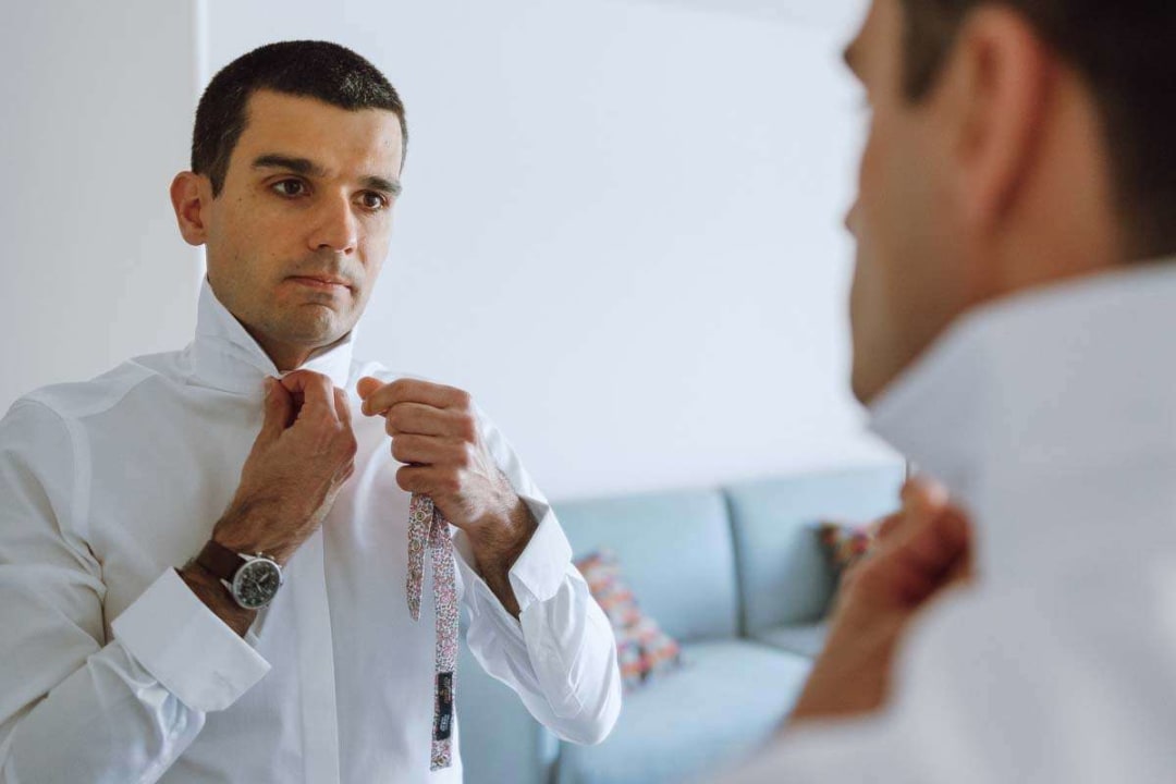 A groom adjusting his tie in front of a mirror before the wedding ceremony.
